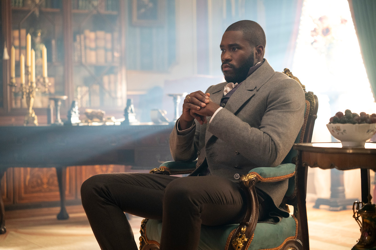 Man sitting thoughtfully in an ornate chair in an elegant, sunlit room with antique furniture, bookshelves, and a lavish table setting, giving a sophisticated and contemplative atmosphere.