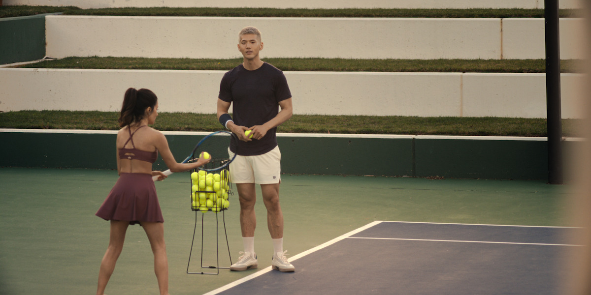 A woman in tennis attire holds a racket while a man stands beside her with tennis balls on an outdoor tennis court, next to a basket of tennis balls, green steps and white walls in the background.