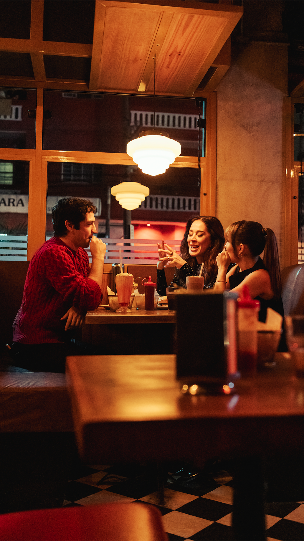 Três pessoas sentadas em uma mesa de bar, com luz baixa e amarela, conversando com bebidas à mesa