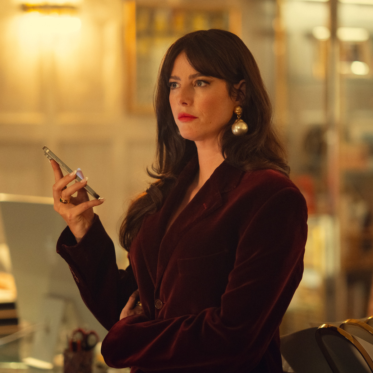 Woman in a burgundy velvet blazer holding a smartphone, standing indoors in a warmly lit office or home with elegant decor, looking focused and thoughtful.