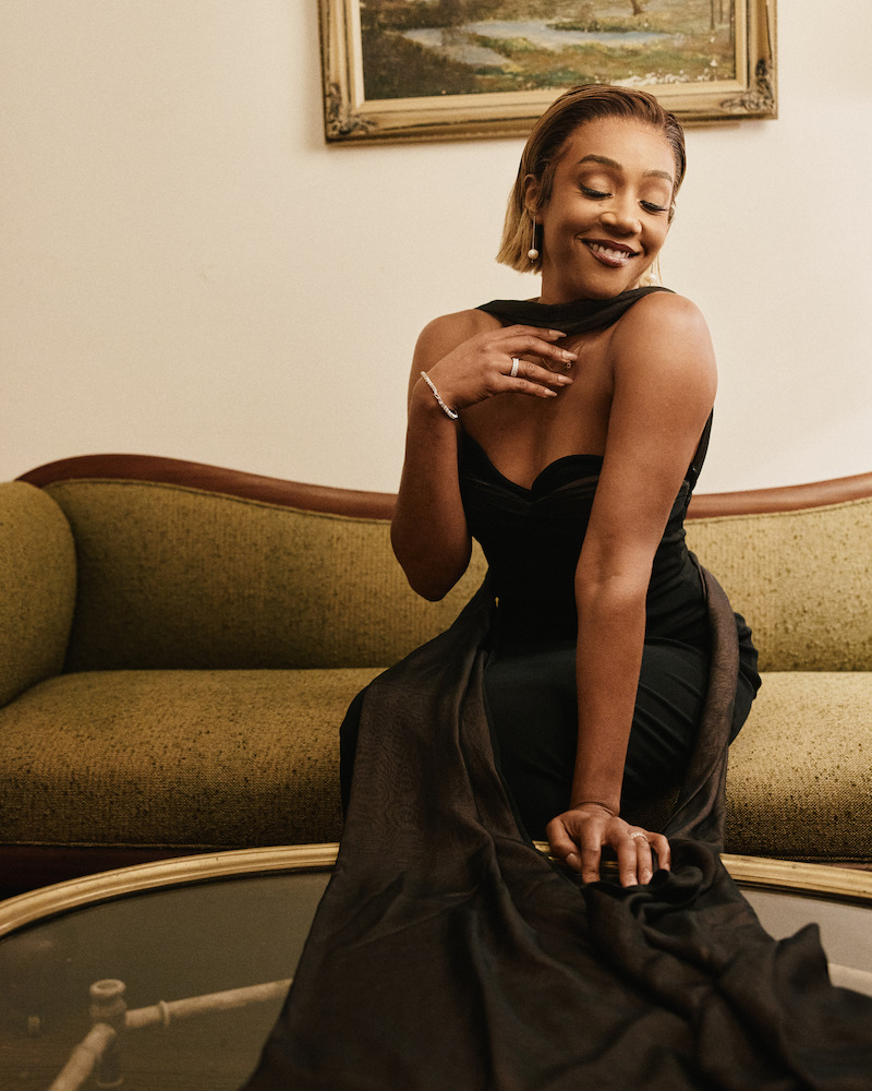 Woman in an elegant black dress sitting on a vintage green sofa in a warmly lit living room, smiling with a hand on her chest, with a painting on the wall and a glass coffee table in front of her.