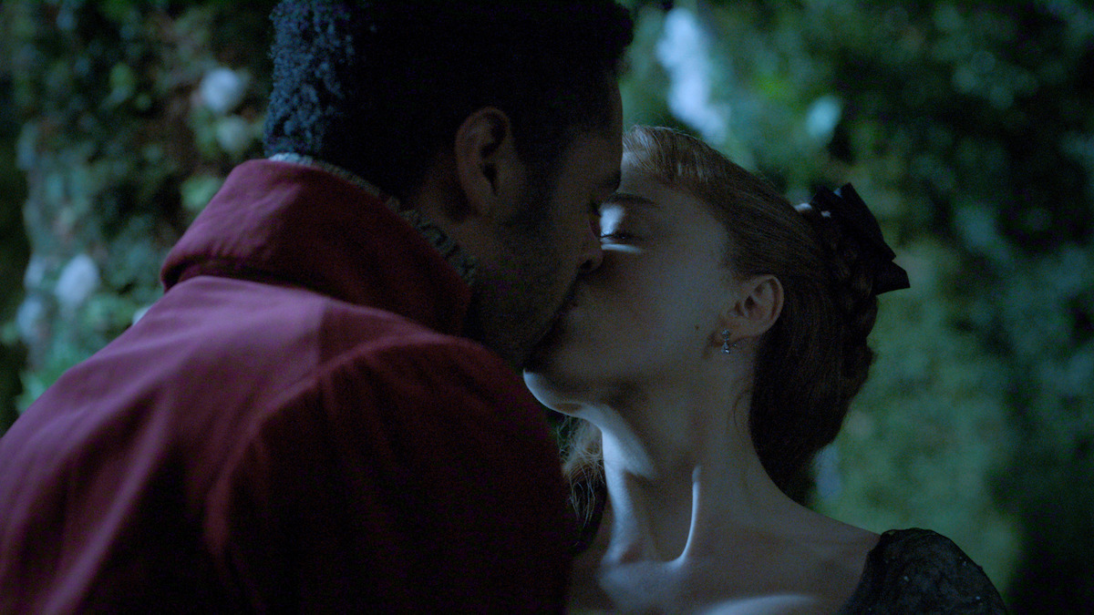 A man and woman dressed in period clothing share a kiss at night in a garden setting, surrounded by greenery and faint moonlight.