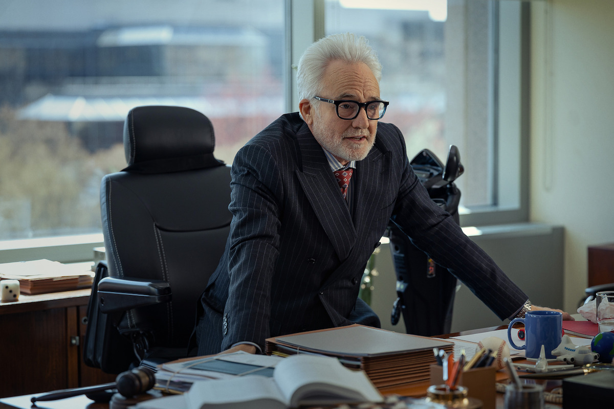 Older man with white hair and glasses, wearing a pinstripe suit, stands leaning on a cluttered desk in a modern office with large windows and cityscape view, appearing engaged or in conversation.