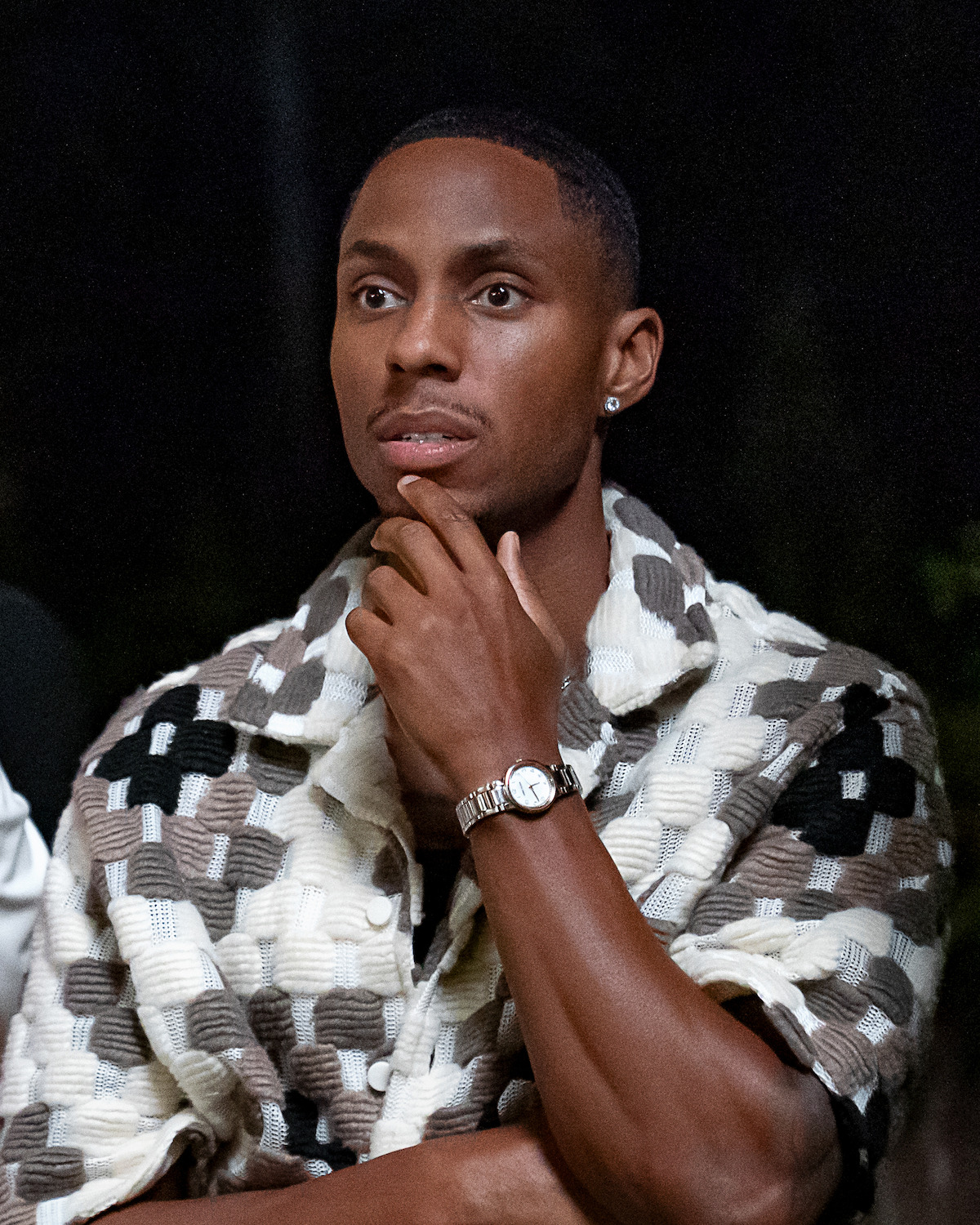 A man sits thoughtfully with his hand on his chin, wearing a patterned jacket and wristwatch, in an indoor or nighttime setting with a dark background.