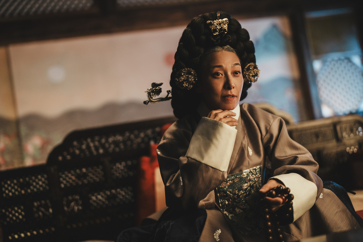 A woman in traditional Korean royal attire with an elaborate hairstyle sits indoors on ornate furniture, holding prayer beads in a dimly lit, historical setting.
