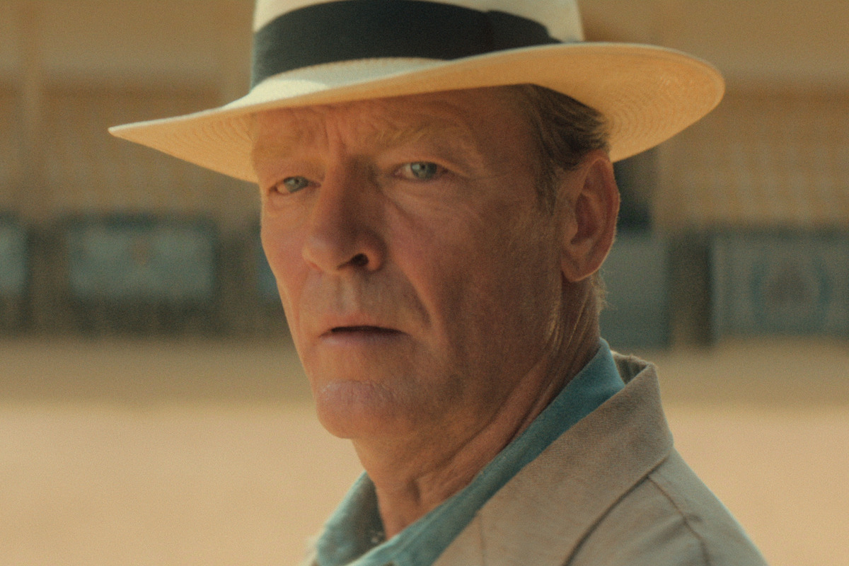 A man in a light suit and white fedora hat stands outdoors in a sunlit arena, looking towards the camera with a serious expression. The background shows empty seating.