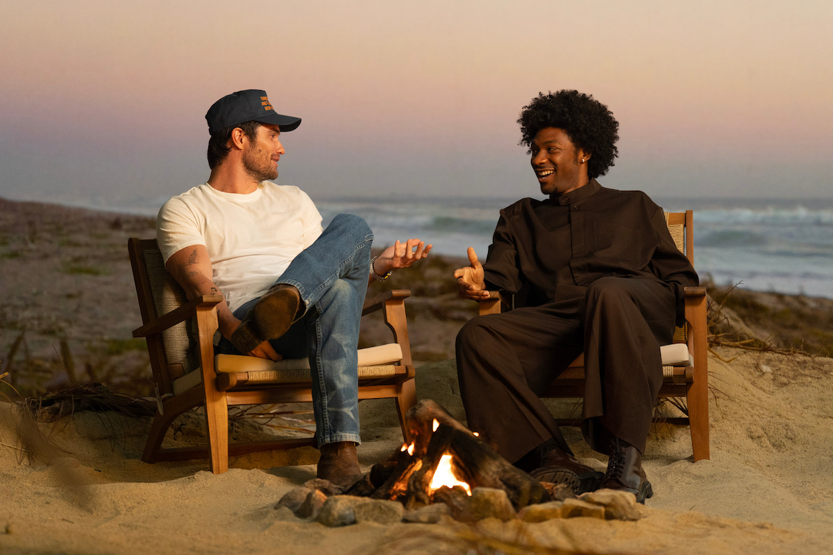 Two people sitting in chairs on a sandy beach by a campfire, talking and gesturing as the sun sets over the ocean in the background, creating a relaxed and friendly outdoor atmosphere.