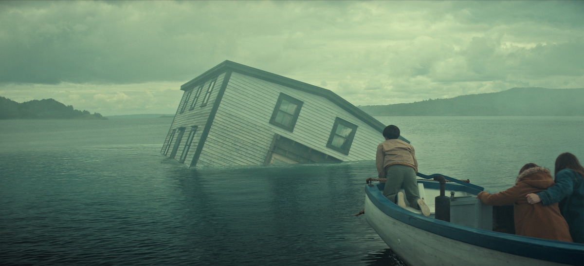 A partially submerged house tilts in a misty lake as three people in a small boat observe, with cloudy skies and distant hills creating a dramatic, mysterious atmosphere.