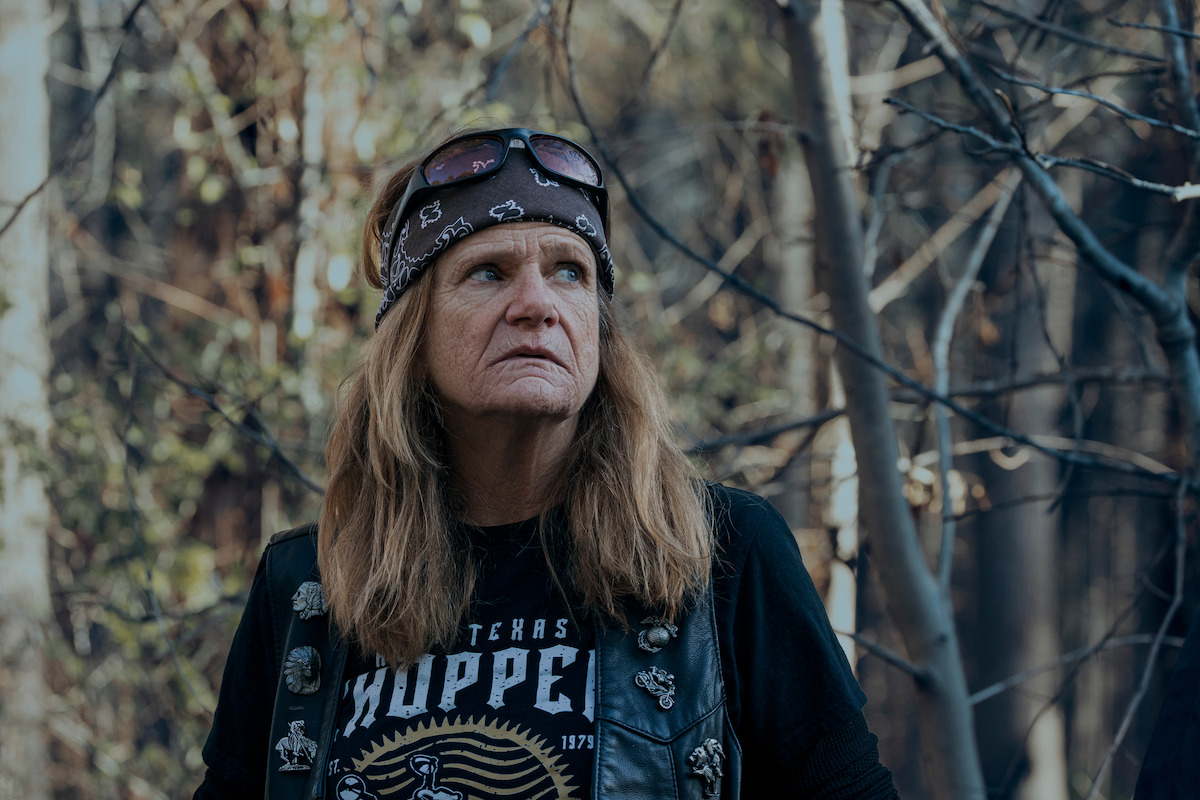 Woman with long hair wearing a bandana, sunglasses, and biker vest stands outdoors in a forest, surrounded by trees and natural light, gazing off to the side with a serious expression.