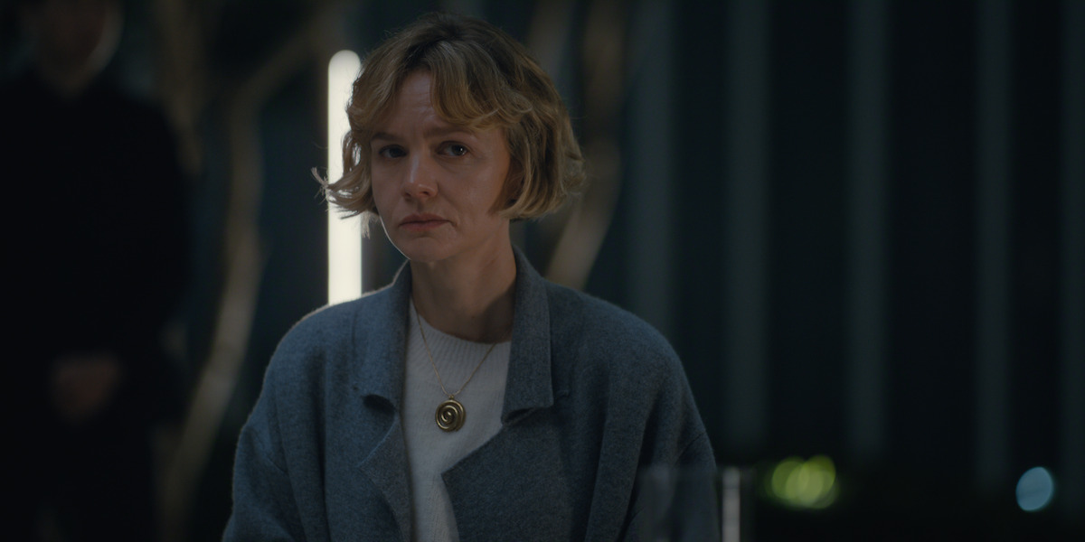 Woman with short blonde hair sits indoors at a table, wearing a gray coat and white shirt with a round pendant necklace, in a dimly lit room with blurred background and vertical light feature.