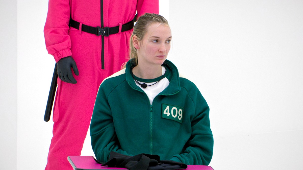 A woman in a green tracksuit with the number 409 sits at a table looking serious, with a guard in a bright pink uniform and black gloves standing behind her in a brightly lit, minimalistic white room.