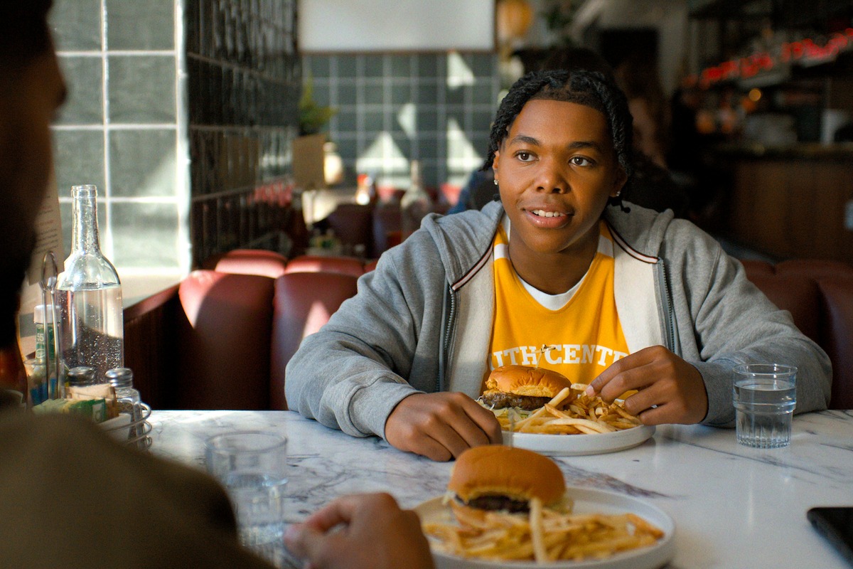 Young person eating a burger and fries in a cozy diner, sitting at a table with someone else, natural light coming through windows, casual and relaxed atmosphere.