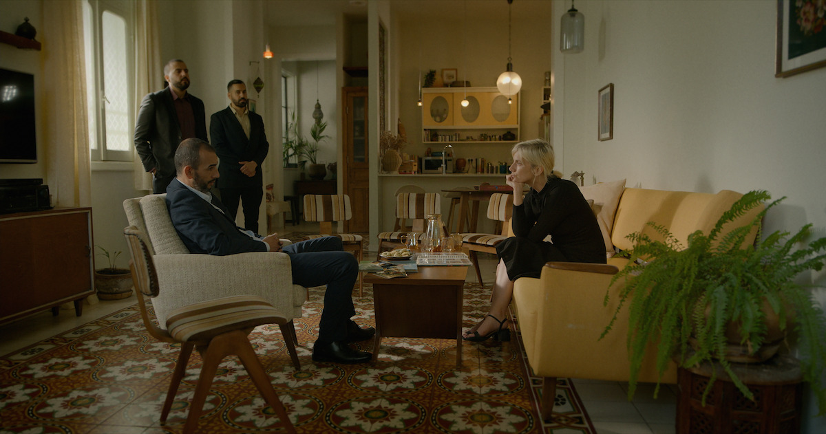 Four people in a living room with mid-century decor; two seated and talking, two standing behind, surrounded by vintage furniture, patterned floor, plants, and warm lighting.
