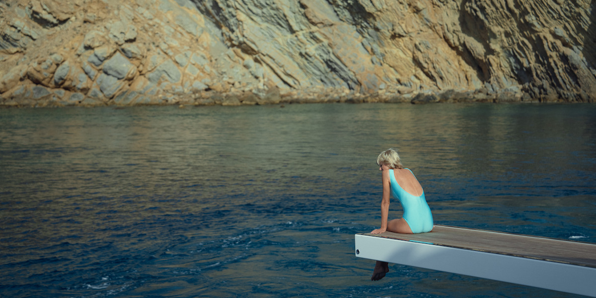 A woman sitting on a diving board in a blue bathing suit looking over the ocean.