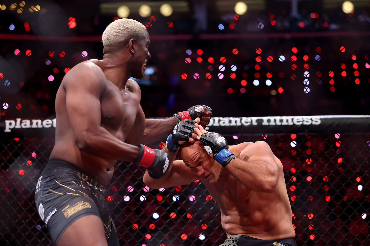 Two male MMA fighters in a cage match, one delivering a punch to the other who is covering up defensively. The arena is filled with bright red and white lights, with "planet fitness" branding visible in the background.