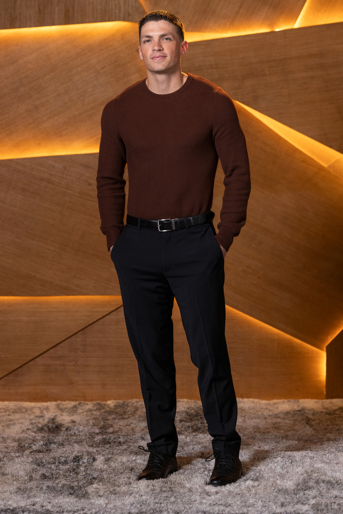 Man standing on a textured rug in front of a modern wooden wall with geometric patterns and warm ambient lighting, wearing a brown sweater, dark pants, and black shoes, posing with hands in pockets.