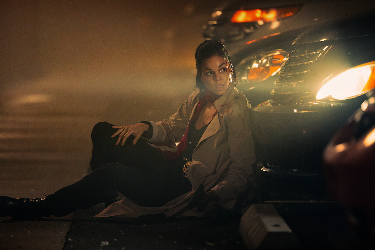 A person in a trench coat sits on the ground at night, leaning against a car with headlights on, in a dark, foggy urban street setting, appearing tense or hiding.