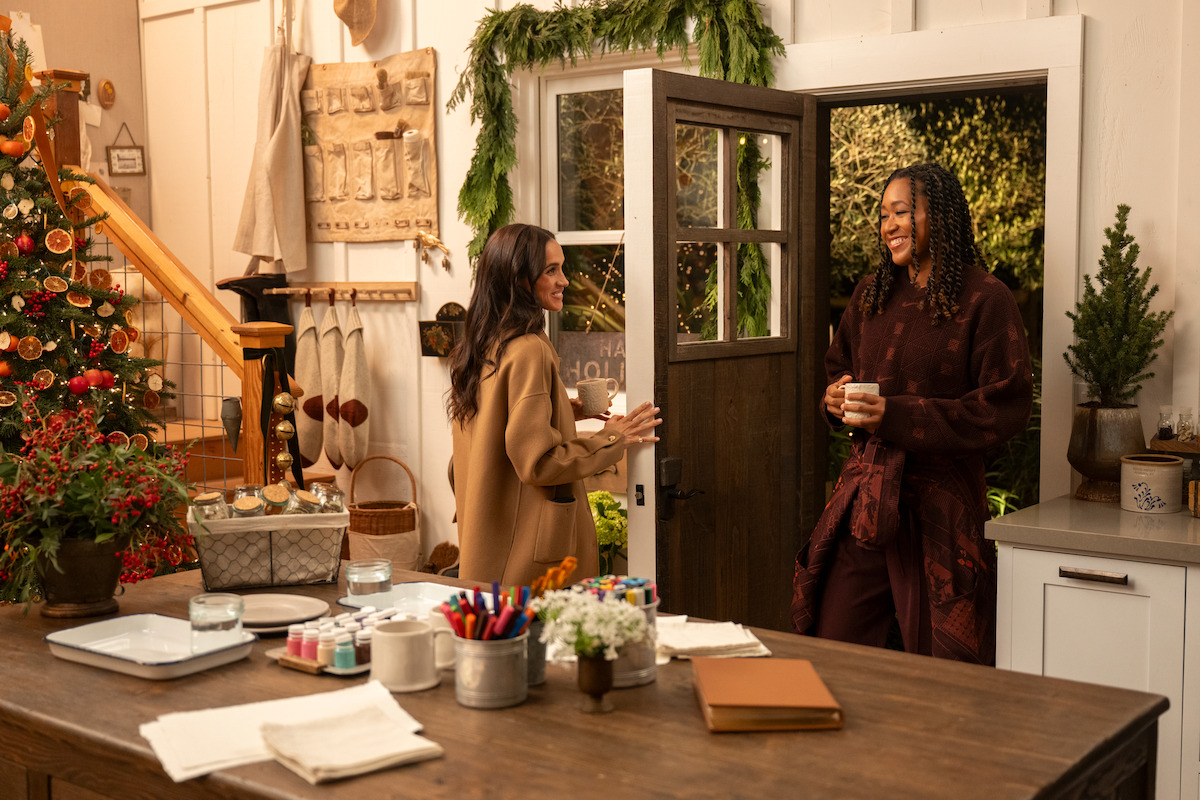 Two women talking and smiling in a cozy, festive kitchen decorated for Christmas with a tree, garlands, crafts, and mugs in hand, creating a warm and inviting holiday atmosphere.