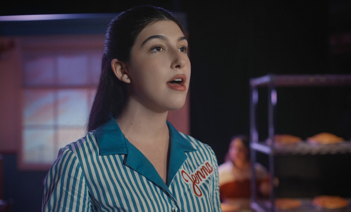 A woman in a blue and white striped uniform with the name "Jenna" sings in a bakery setting, with metal racks of pies and a blurred figure in the background.