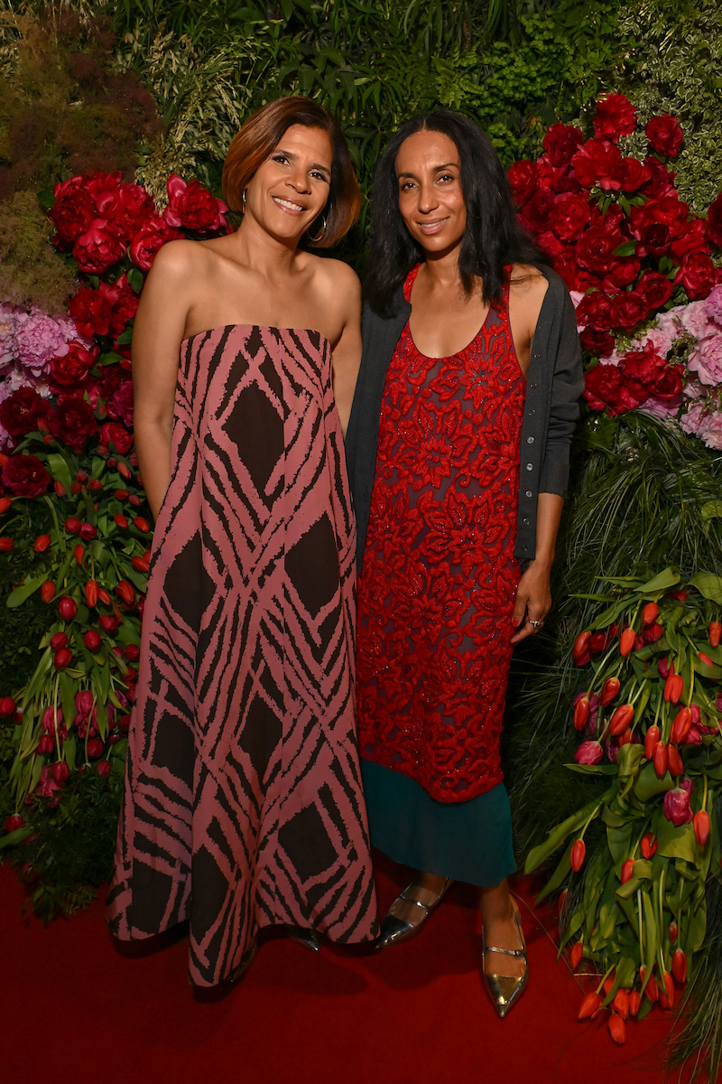 Two women pose together on a red carpet, wearing patterned dresses, surrounded by lush greenery and vibrant red and pink flowers in an elegant indoor garden setting.