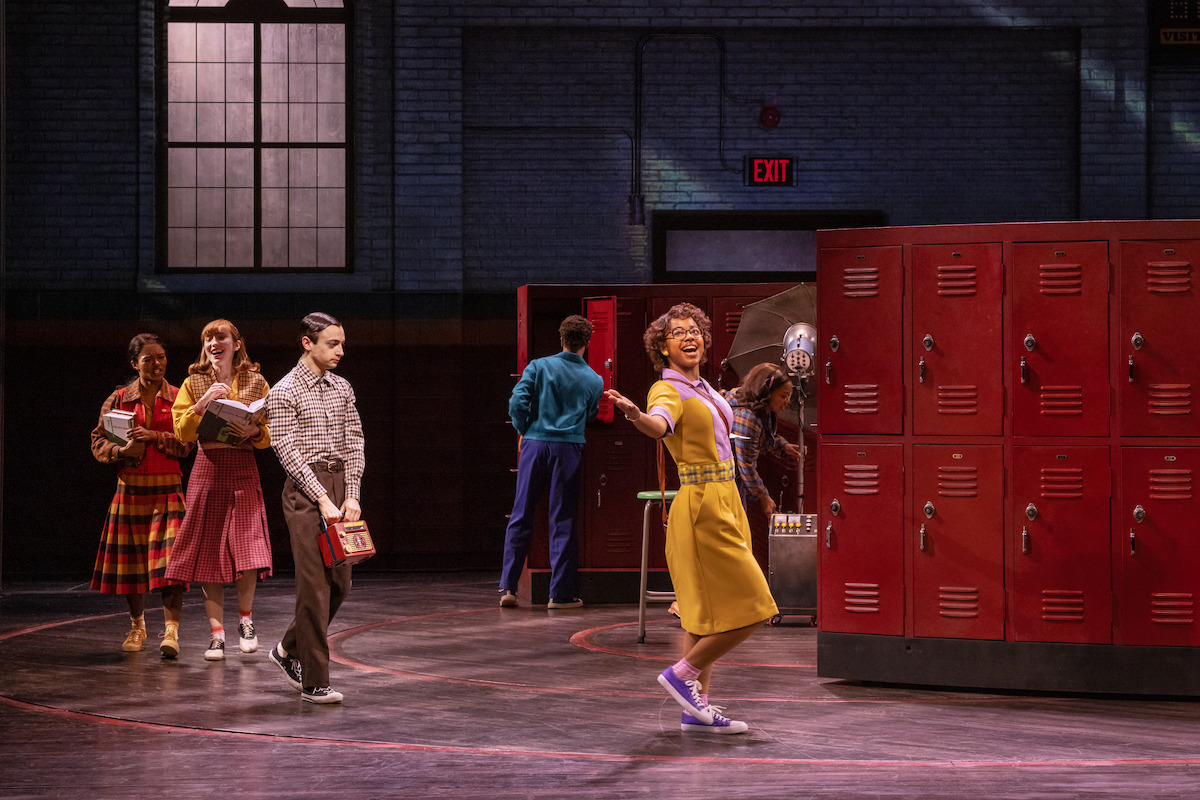 Several actors in 1950s-style clothing perform in a high school hallway set with red lockers and brick walls, some reading or carrying books, one in a yellow dress joyfully singing or dancing center stage.