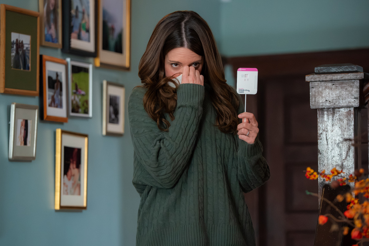 Woman in green sweater stands indoors near staircase and wall with framed photos, holding a Covid test while partially covering her face, autumn decorations visible in foreground.