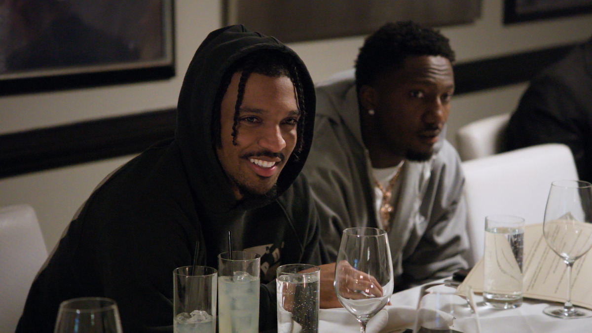 Two men sit at a restaurant table set with glasses, menus, and water. One man, wearing a hoodie, smiles while conversing. The setting is casual and relaxed with contemporary decor.