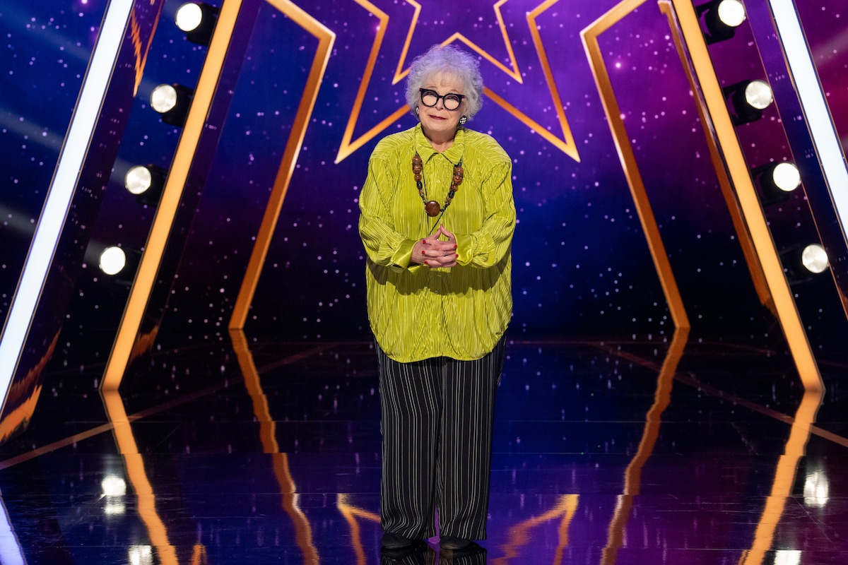 Susan Rice, con cabello blanco y gafas, se encuentra en un escenario brillante con luces brillantes, vestida con una blusa amarilla, pantalones a rayas y un collar. El fondo presenta una gran estrella brillante y un patrón estrellado de color púrpura.
