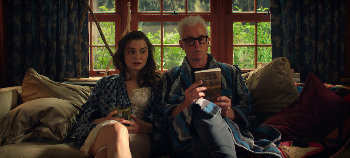 Rachel Weisz, who plays M., and John Slattery, who plays John, are wearing robes and sitting huddled on a cozy couch in a warmly lit living room with windows and greenery outside. One person is holding a mug while the other is reading a book surrounded by pillows and patterned curtains.