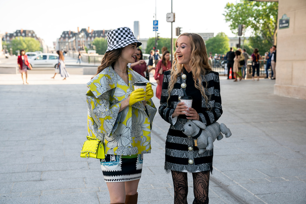 Emily and Camille drinking to-go coffees. Emily is wearing some sort of checkerboard-pattern bucket hat?