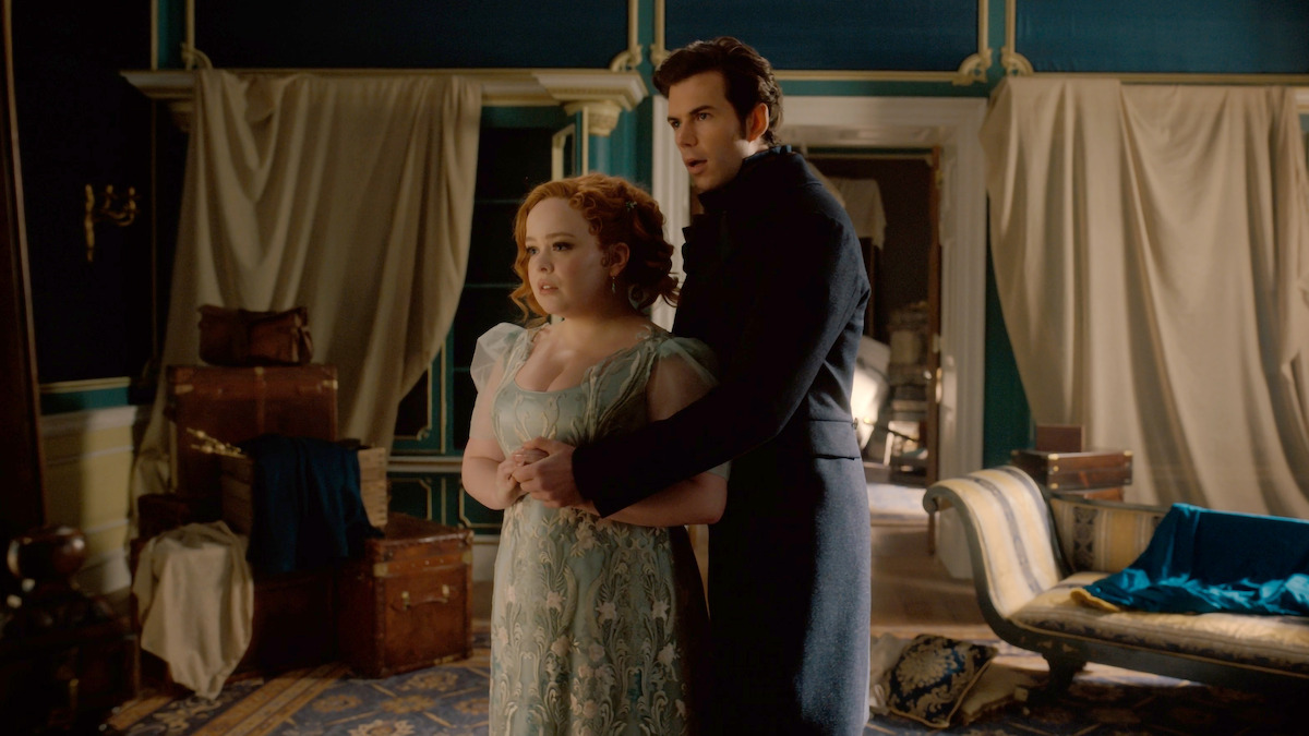 A man and woman in period costumes stand close together in an ornate, dimly lit room with vintage furniture, trunks, and draped curtains, suggesting a dramatic or emotional moment in a historical setting.