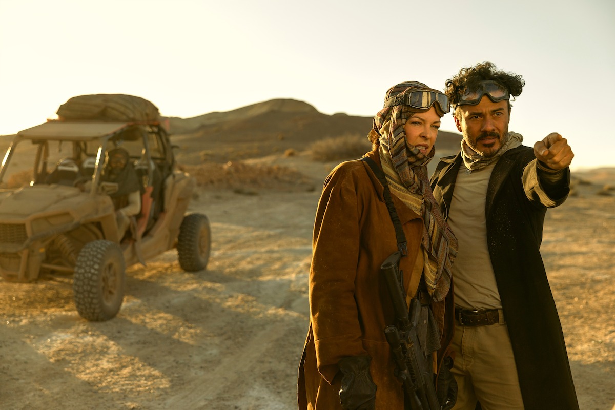 Two people in rugged, post-apocalyptic clothing and goggles stand in a sunlit desert next to an off-road vehicle, with mountains in the background. One person points into the distance, suggesting urgency or direction.