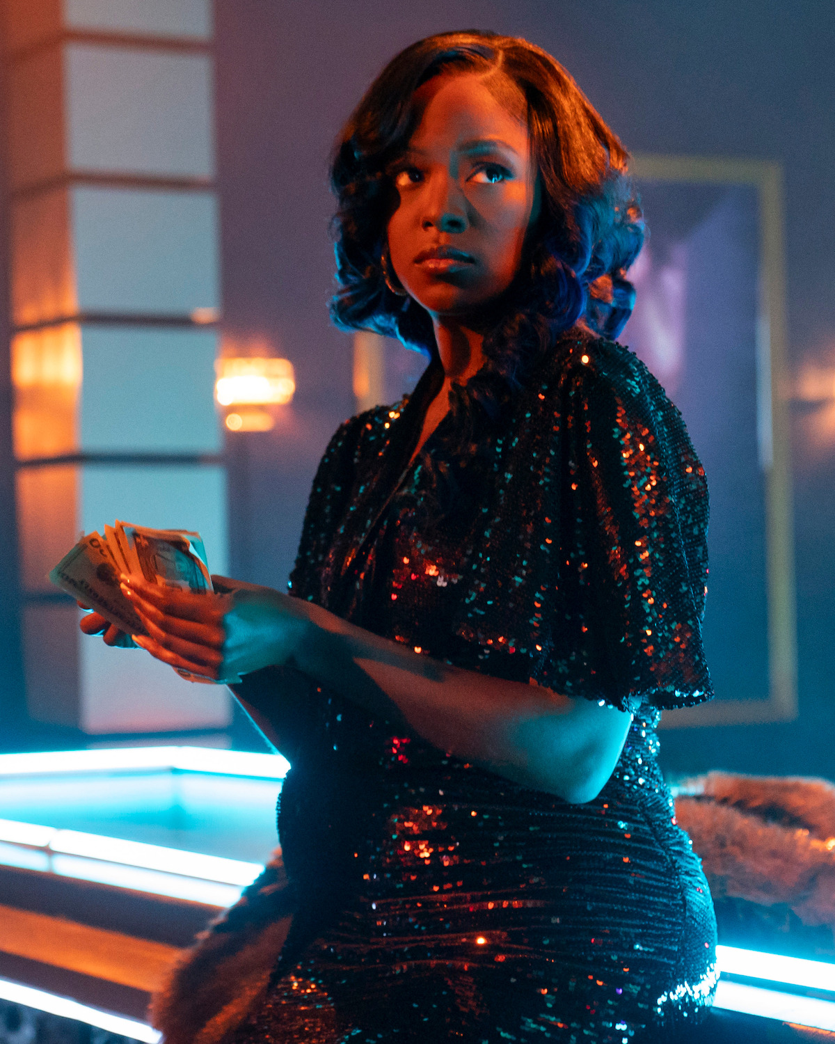 Woman in a sparkly black dress holding cash, standing by a bar in a dimly lit, glamorous nightclub with blue and orange lighting and modern decor.