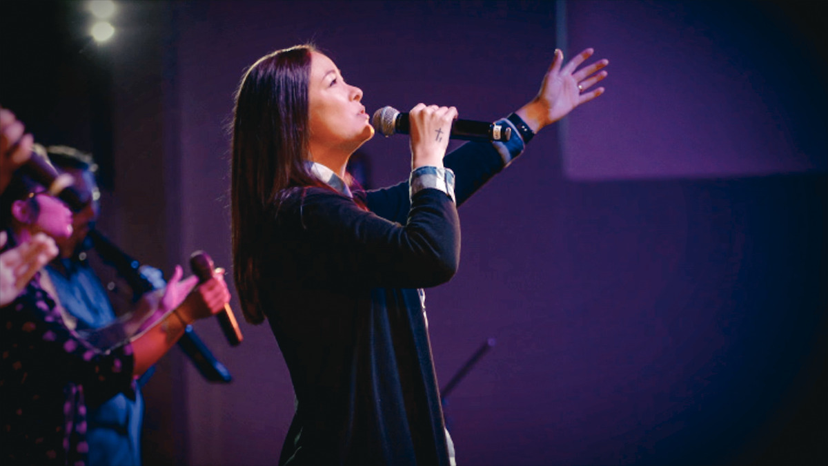 Woman singing passionately into a microphone on stage, with colorful stage lighting and other musicians visible in the background, suggesting a live music or worship performance setting.