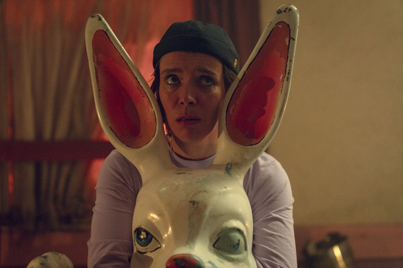 Person wearing a black cap and light shirt sits behind a large white rabbit statue with pink ears in a dimly lit room with beige curtains and warm lighting.