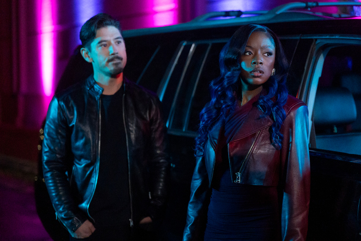 Two people in leather jackets stand by a car at night, illuminated by dramatic pink and blue lighting, creating a tense and mysterious mood in an urban environment.