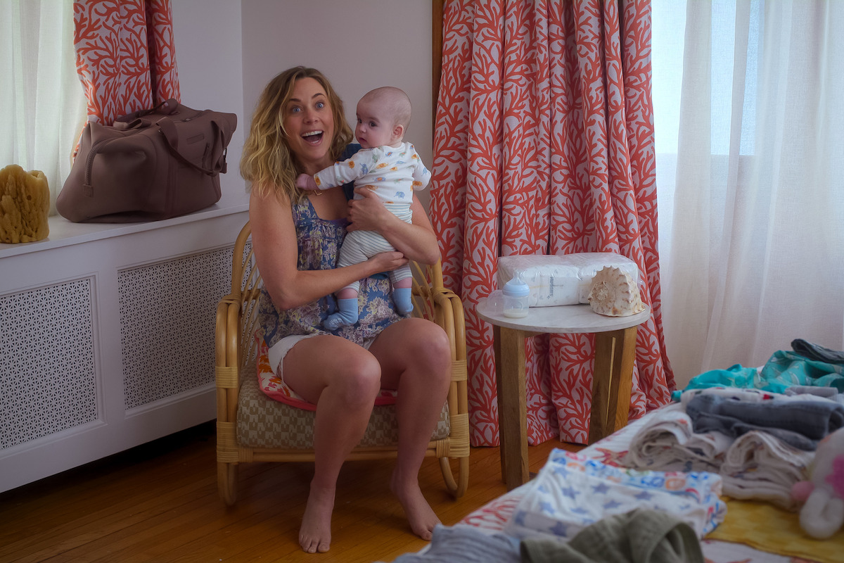 A smiling woman sits on a wicker chair holding a baby in a bright room with patterned curtains, a small side table, and laundry or baby clothes spread out on a bed nearby.