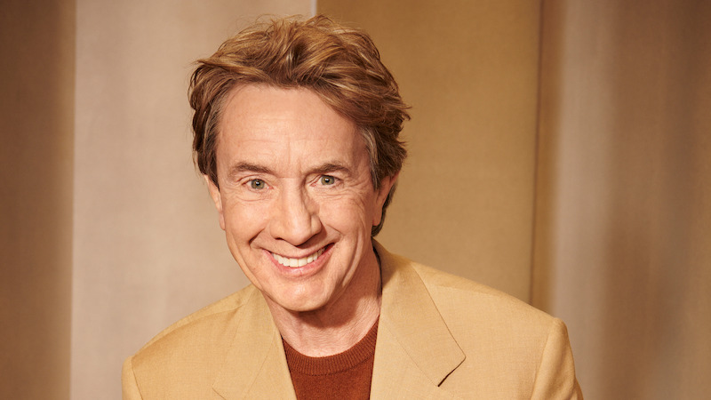 Martin Short smiles at the camera wearing a tan jacket.