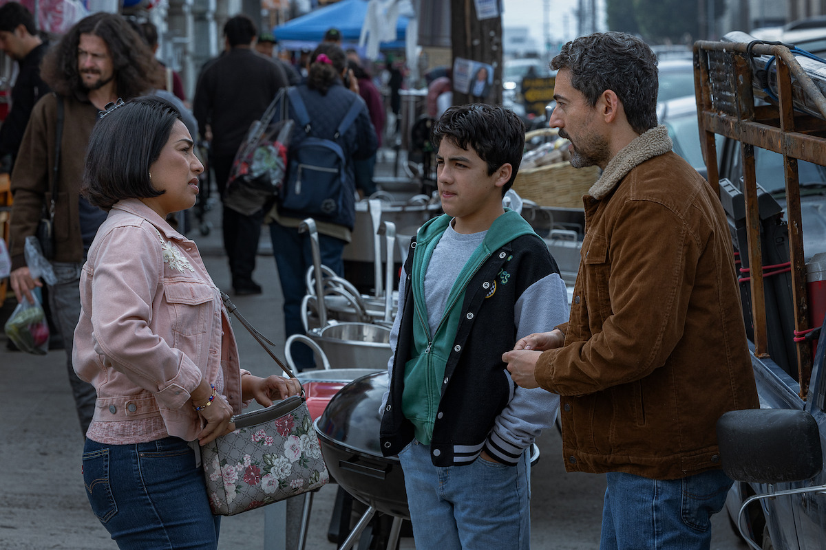 A woman, a boy, and a man stand talking on a busy outdoor market street with vendors, tables, and people in the background.