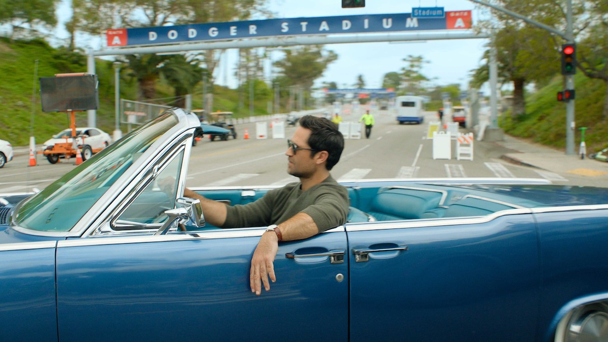 Man driving a blue convertible near Dodger Stadium entrance on a sunny day, relaxed atmosphere, street scene with greenery, traffic lights, and stadium sign in the background.