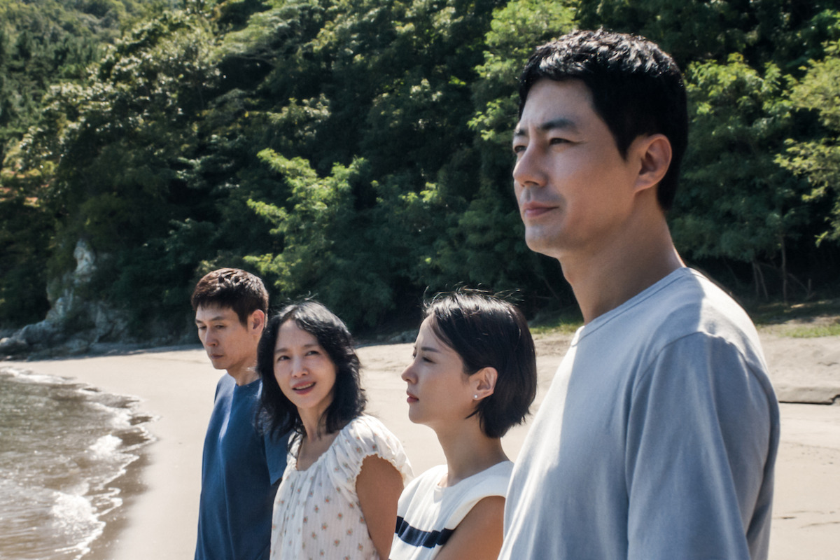 Four people stand side by side on a sandy beach, facing the water, with green trees and sunlight in the background. They appear thoughtful and relaxed, enjoying a peaceful day at the shoreline.