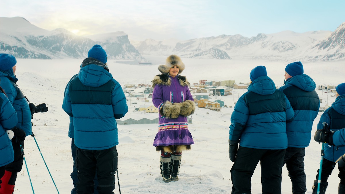 Mulher sorridente, com roupas tradicionais de acabamento em pele, em uma paisagem ártica coberta de neve. Ao redor dela, cinco pessoas de jaqueta de inverno azul. Ao fundo, um vilarejo cercado de montanhas, sob um céu claro.