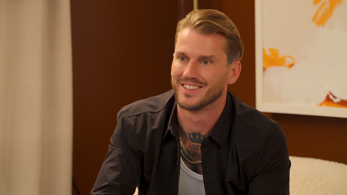 Smiling man with blond hair and visible chest tattoos wearing a black shirt sits indoors on a chair, with warm brown walls, a curtain, and abstract art in the background.