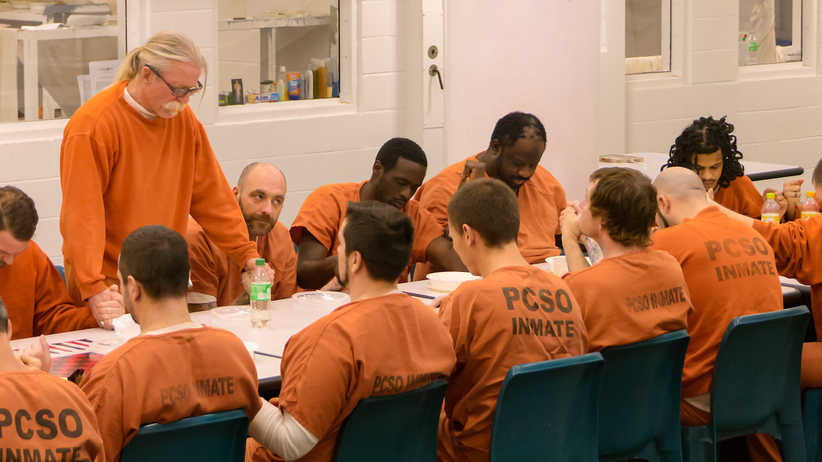 Group of inmates in orange uniforms seated at tables in a prison common area, some interacting with a standing man, with institutional white walls and windows in the background.