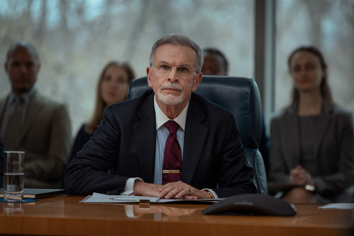 Older man in suit sits at conference table with documents and microphone, appearing to listen or speak during a formal business meeting, with other people seated in the background in a modern, professional office setting.