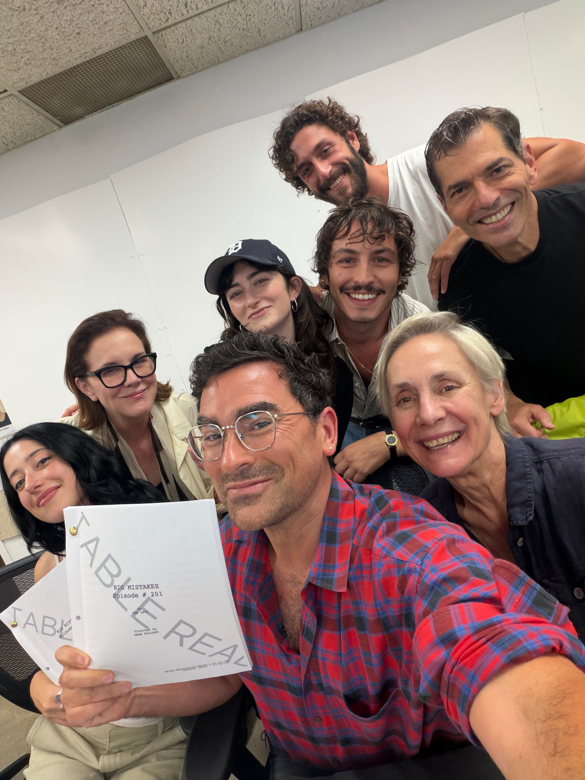 Group of eight people smiling and gathered indoors, holding scripts in a bright classroom or office setting, under fluorescent ceiling lights, suggesting a casual or creative meeting or script reading session.