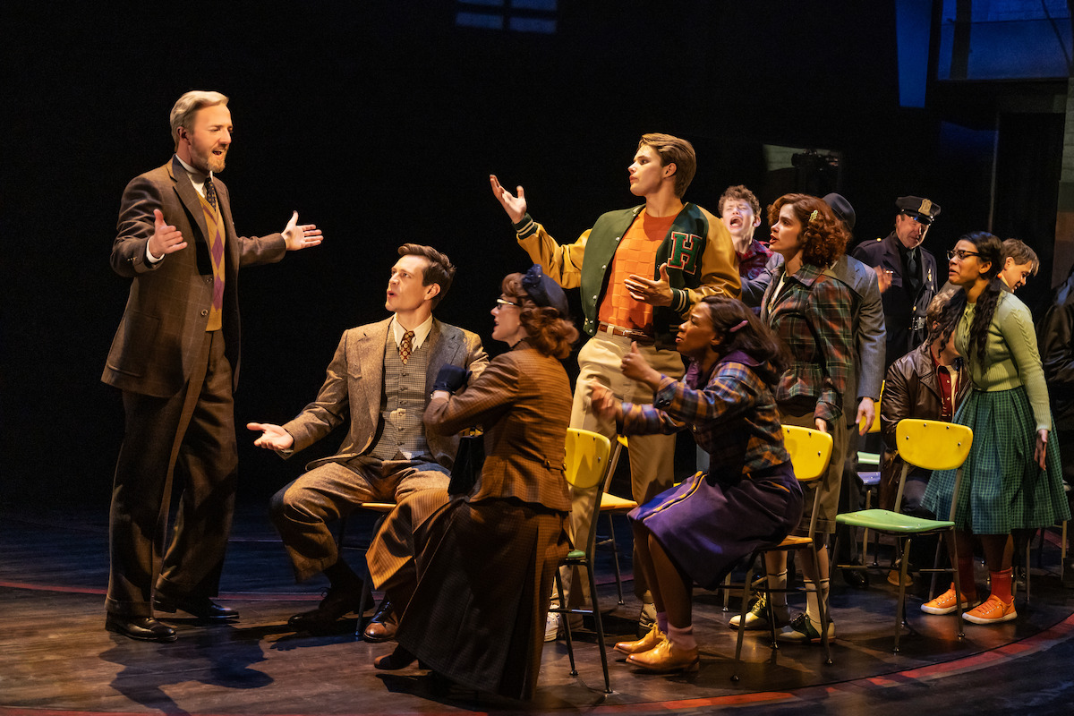 A group of actors on stage performing a dramatic scene, with one man standing and gesturing as others sit or stand around him in period costumes, set in a theater with dim lighting and classroom chairs.