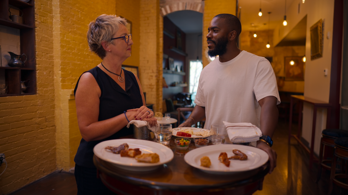 Two people stand by a round table with plates of food, talking in a warmly lit restaurant or café with yellow brick walls and modern decor, surrounded by dining tables and hanging lights.