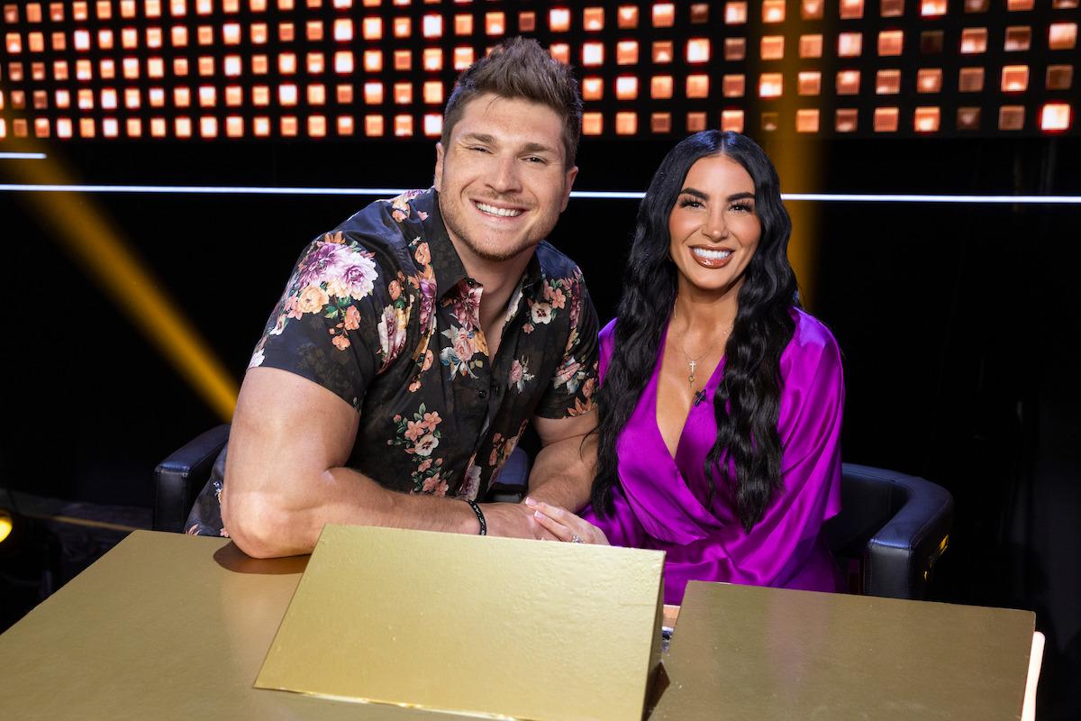 A smiling man in a floral shirt and a woman in a purple outfit sit together at a golden table on a brightly lit stage, creating a cheerful and celebratory mood.