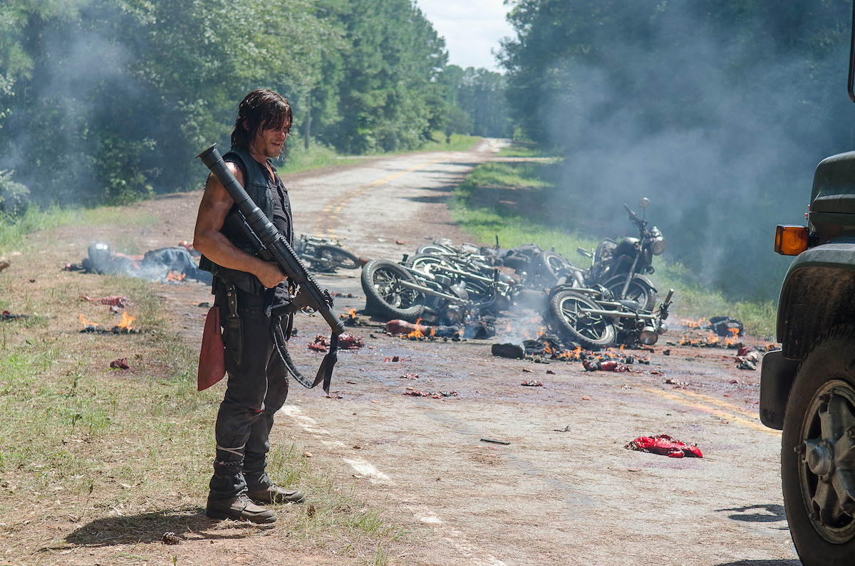 Daryl holds a rocket launcher in front of several burning motorcycles. 
